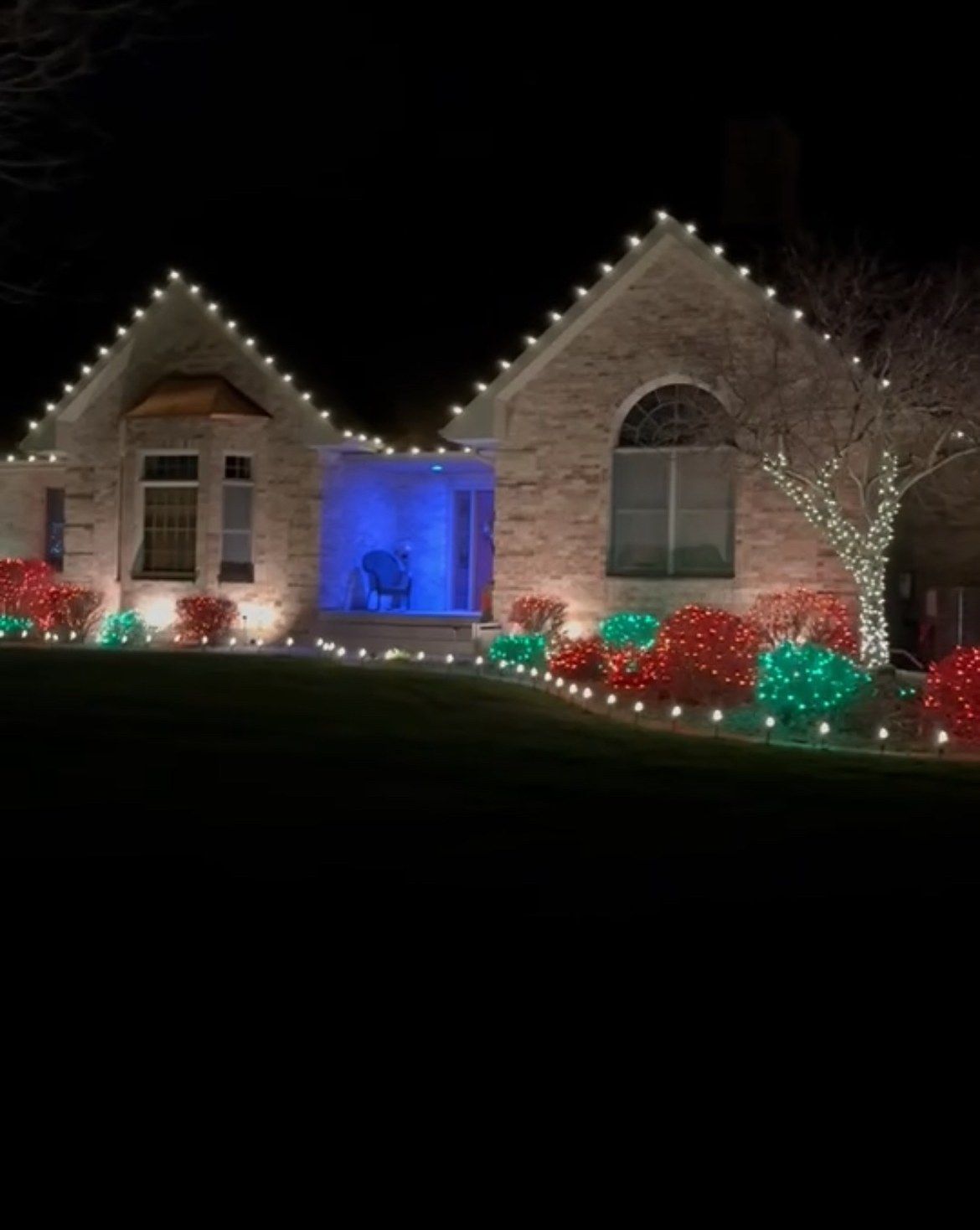 Brick ranch with warm white roofline lights, red and green netted shrubs and a wrapped front-yard tree