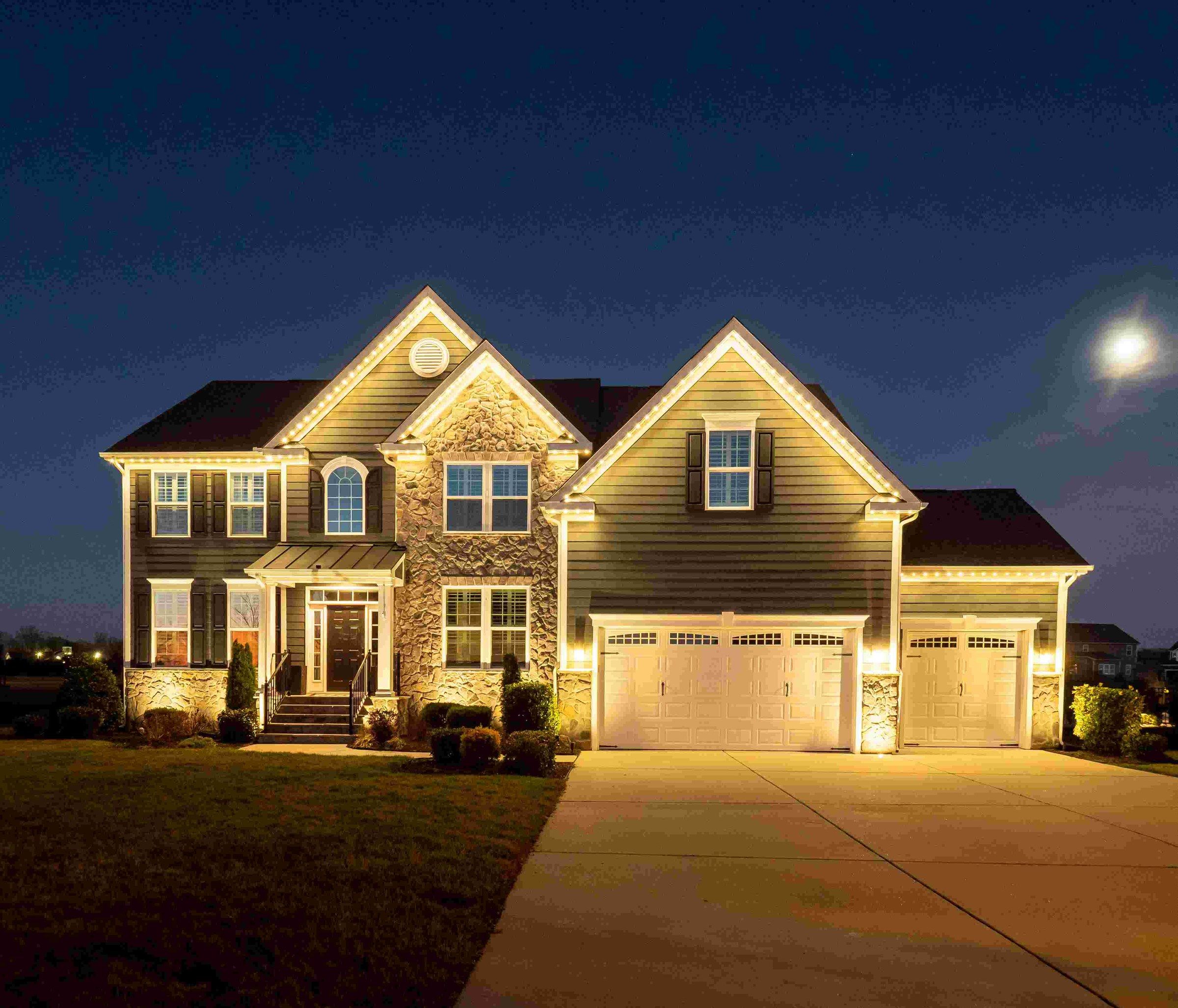 Large stone-accented two-story with warm white lights on every roofline and dormer under a moonlit sky