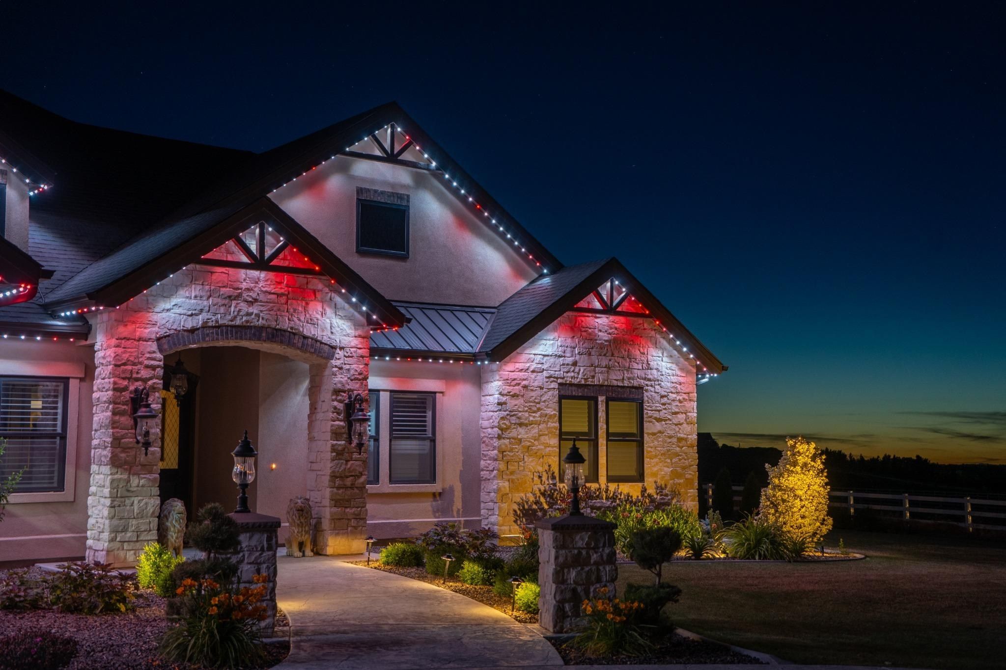 Stone and stucco home with permanent lights in red and white along the gables and a lit evergreen